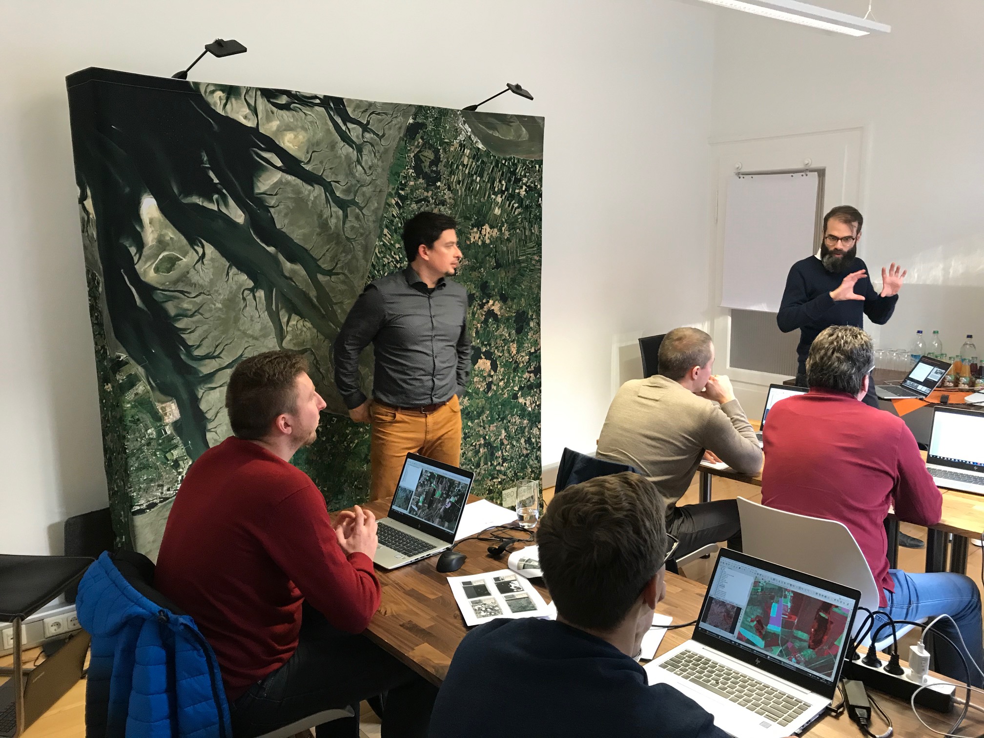 LAAS QGIS Training: People sitting behind laptops in a room, one standing at the edge in front of a large satellite image. Someone is presenting at the front.